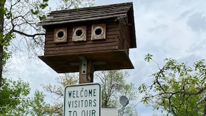 A wooden sign with a welcome message for a country retreat, a wooden birdhouse, and a white house with a porch. - A  peaceful country retreat a block away from the beach,with beautiful sunsets.  (Dunnottar)