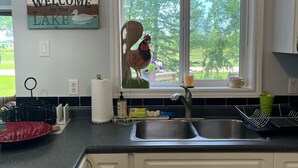 A kitchen with a sink, a window with a view of trees, a welcome sign, and a decorative rooster. - A  peaceful country retreat a block away from the beach,with beautiful sunsets.  (Dunnottar)
