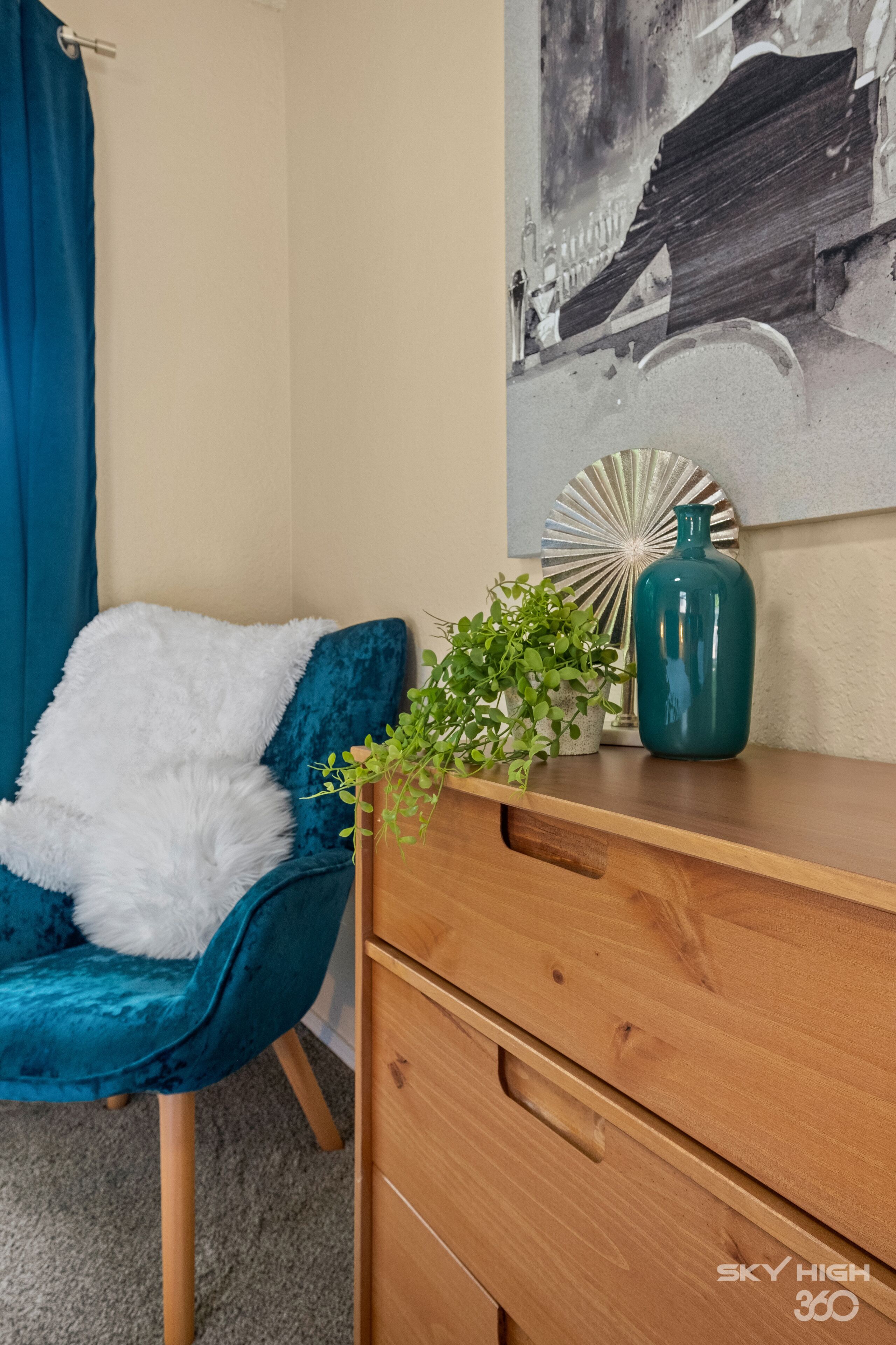 Un fauteuil de couleur turquoise agrémenté d’un plaid blanc et moelleux, une commode en bois avec des tiroirs, un vase turquoise contenant une plante, et un mur orné d’un miroir en forme de soleil et d’un tableau en noir et blanc.