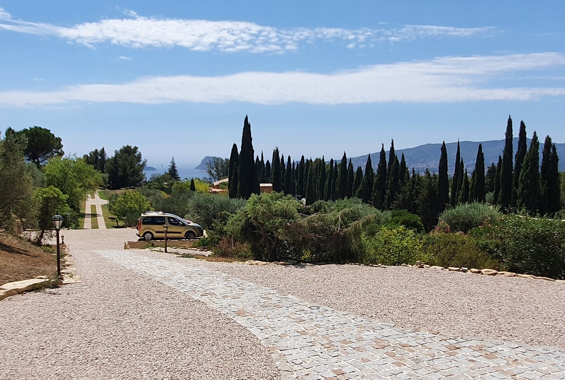 Un sentier pavé mène à un monument en forme de croix, entouré de cyprès et dominé par un paysage montagneux.