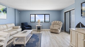 Una sala de estar con un sofá color beige, un sillón azul, una mesa de centro blanca y un tapete azul. Se ve una ventana con vista al mar, una lámpara sobre una mesita y un aparador con un barco modelo.