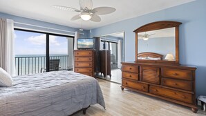Un dormitorio con cama, cómoda y ventilador de techo. Se ve el océano a través de un ventanal.