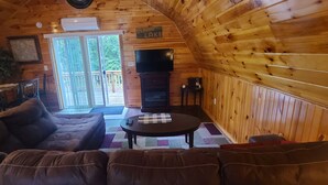 A cozy cabin interior with a large couch, a small table, a fireplace, and a sliding glass door leading to an outdoor deck. - Cute romantic studio  (Moultonborough)
