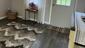 Une pièce avec un parquet en bois foncé, un tapis à motifs, un panier en osier, une petite table avec un vase, une porte avec une fenêtre et un luminaire.