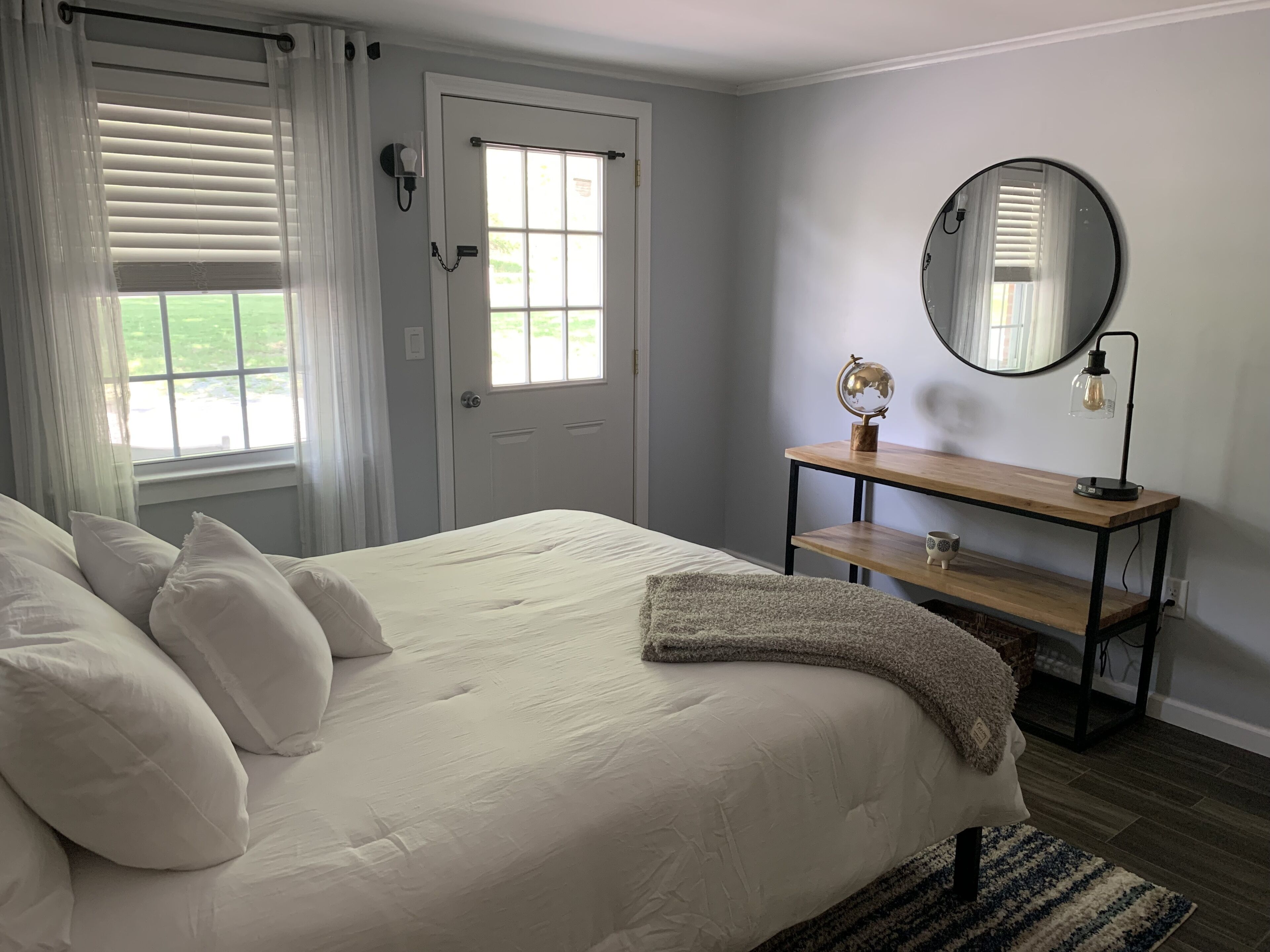 Un dormitorio con cama, una mesita de noche de madera, un espejo redondo y una ventana con persianas.