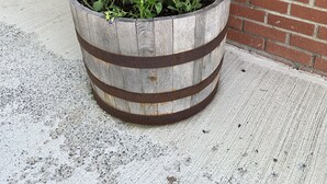 Un grand pot en forme de tonneau en bois, contenant un arbre à feuilles persistantes et des fleurs, est placé sur une surface en béton devant un bâtiment en briques doté d’une fenêtre et d’une enseigne indiquant « BIENVENUE ».