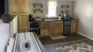 Une cuisine compacte avec des meubles en bois, un four à micro-ondes, une machine à café et un grille-pain. Des étagères au-dessus de l’évier servent à ranger les tasses et les verres.