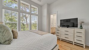 Een slaapkamer met een groot bed, een televisie op een dressoir en uitzicht op bomen door de ramen.