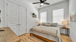 Um quarto com cama, mesas de cabeceira, ventilador de teto e janela com persianas.