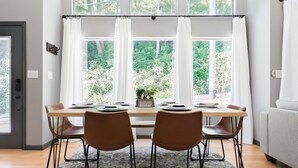 Un comedor con mesa dispuesta para cuatro personas, ventanas amplias con cortinas y vistas al exterior con vegetación.