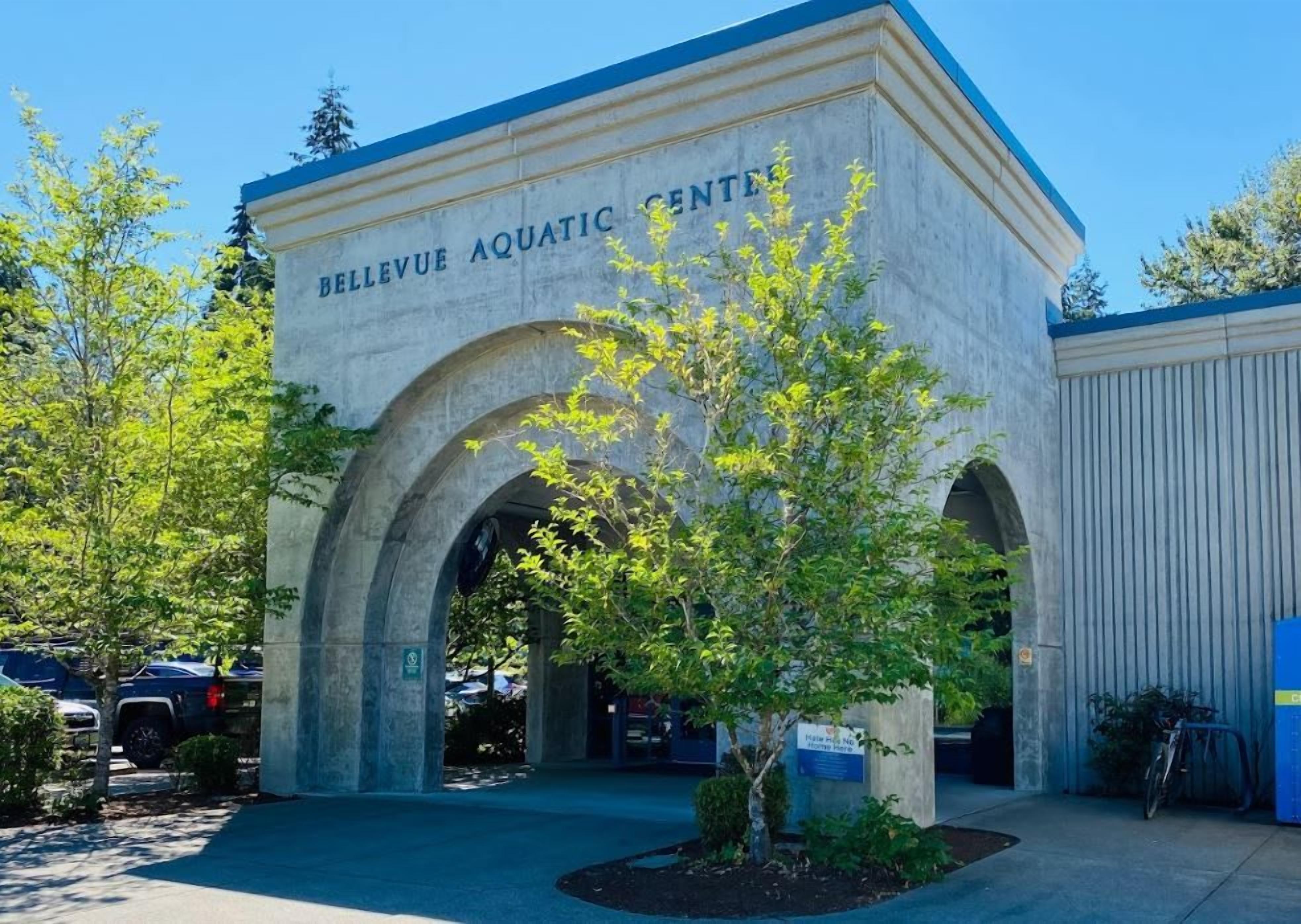 Un bâtiment portant l’inscription « CENTRE AQUATIQUE DE BELLEVUE », avec une entrée en arc de cercle, un petit arbre au premier plan et des voitures garées à l’arrière-plan.