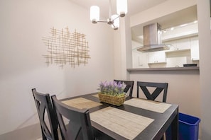 A dining area with a dark wooden table, black chairs, and a woven placemat with purple flowers. A modern light fixture hangs above, and a kitchen is visible in the background. - Modern Microsoft Redmond Campus Condo with Balcony (Bellevue)