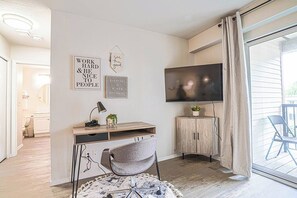Un salon moderne comprenant un bureau, une chaise, un téléviseur et une porte-fenêtre coulissante donnant sur un balcon.