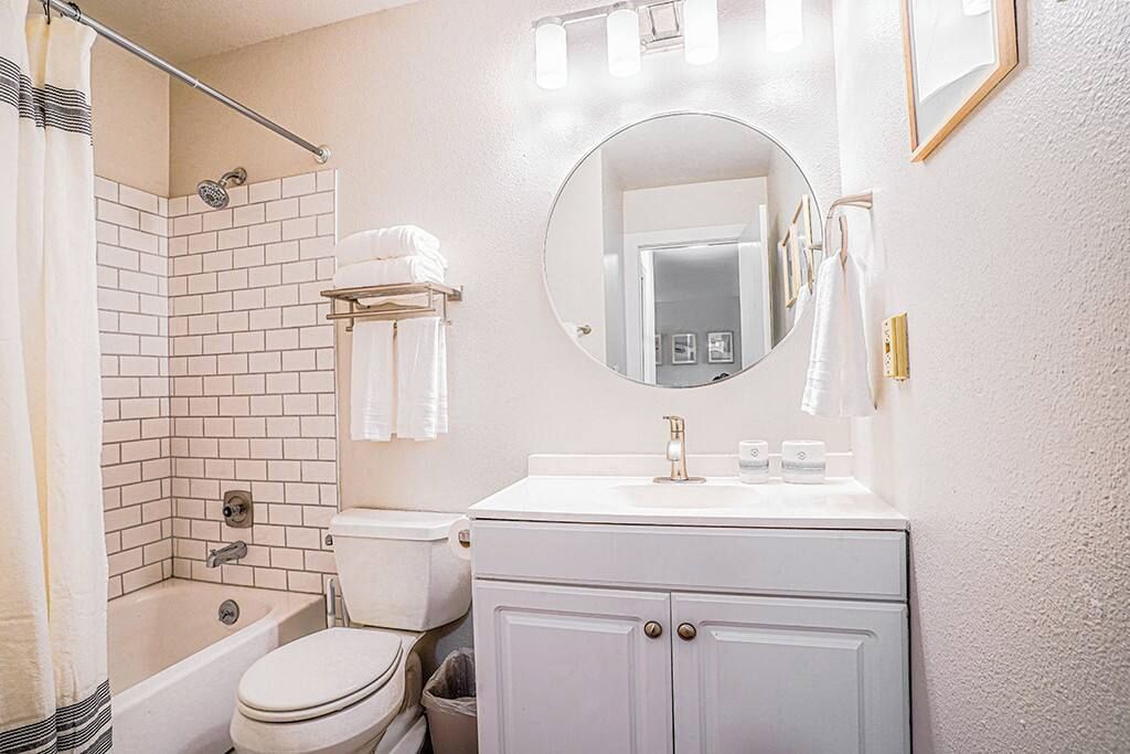 A bathroom with a white bathtub, toilet, and sink, a round mirror, and a shower area.