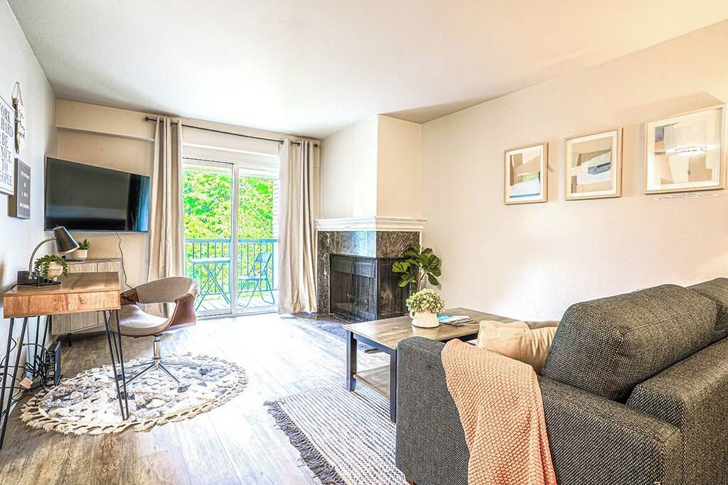 A living room with a sofa, a coffee table, a fireplace, a TV mounted on the wall, and a balcony with curtains.