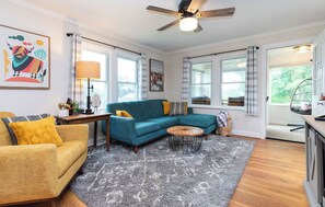 Una sala de estar moderna con un sofá seccional color turquesa, un sillón amarillo y una mesa de centro de madera. Hay un ventilador de techo, ventanas amplias con cortinas y una alfombra gris en el suelo.