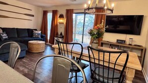 Un salon moderne avec un canapé bleu foncé, une table à manger en bois avec des chaises, un téléviseur fixé au mur et une grande fenêtre avec des rideaux.