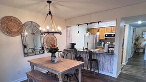 Un coin repas avec une table et un banc en bois, une cuisine équipée d’appareils modernes et un couloir menant aux autres pièces.