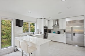 Una cocina moderna con armarios blancos, una isla grande, electrodomésticos de acero inoxidable y vistas a árboles a través de las ventanas.