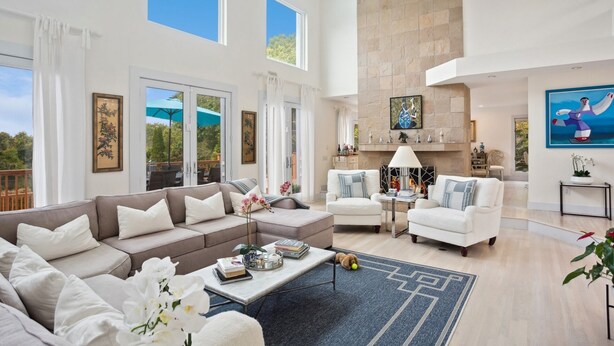 A spacious living room with a high ceiling, large windows, and a fireplace.