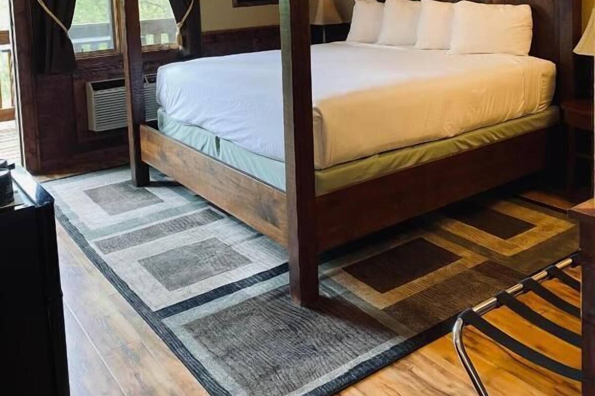Una cama con dosel de madera y ropa blanca, una mesita de noche con lámpara, y vistas a zonas verdes por la ventana.