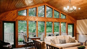 A cozy cabin interior with a large wooden ceiling, a sofa with cushions, a dining table, and large windows offering a view of the outdoors. - Sylvan Valley Lodge & Cellars (Sautee Nacoochee)