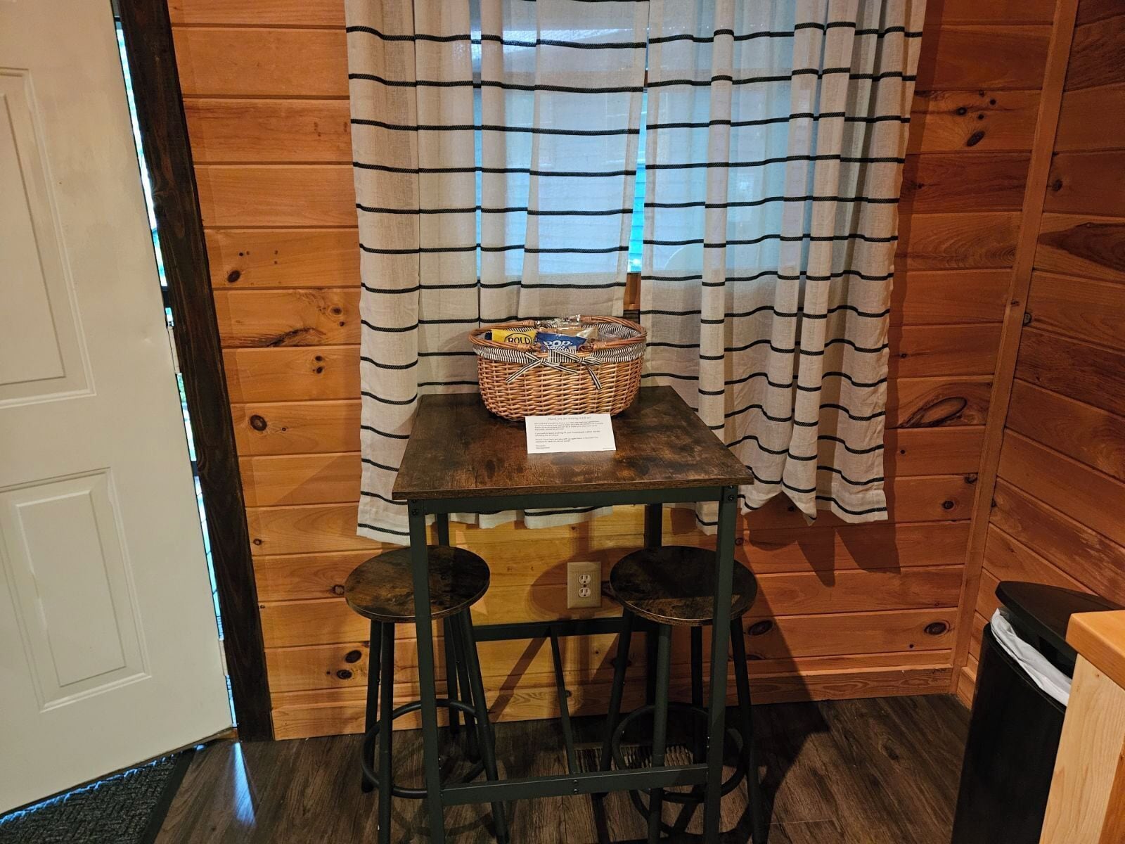 Una pequeña habitación de madera con una mesa, dos taburetes, una cesta sobre la mesa y una cortina a rayas.