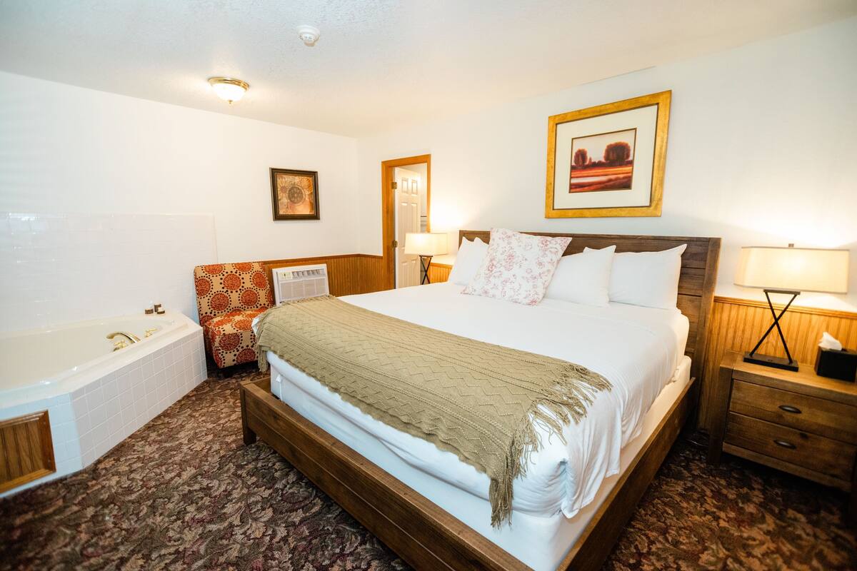 Habitación de hotel con una cama grande, una bañera, un cabecero de madera, una mesita de noche y un cuadro enmarcado en la pared.