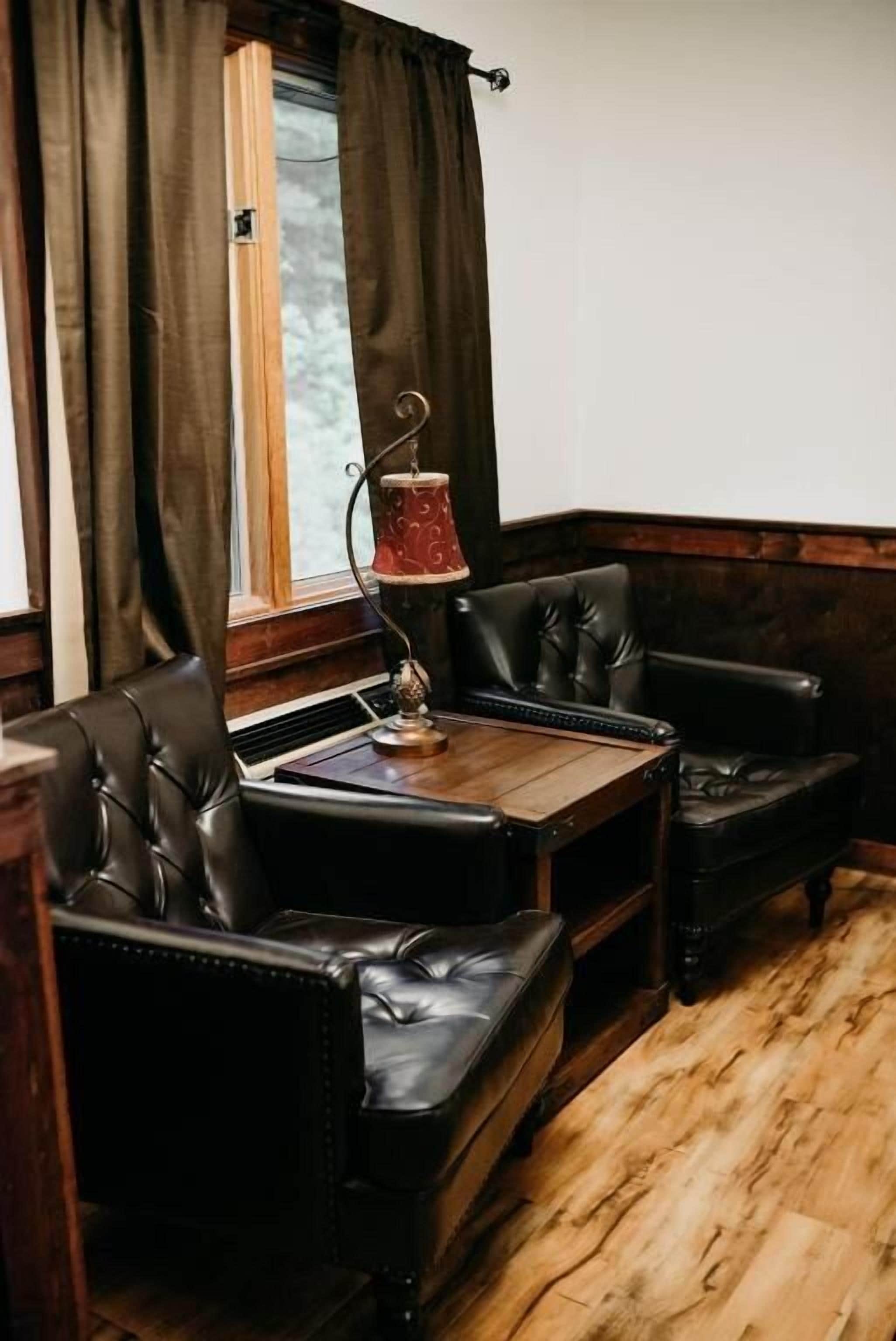 Una habitación con piso de madera, sofá de cuero marrón oscuro, una mesita de madera y una ventana con cortinas.