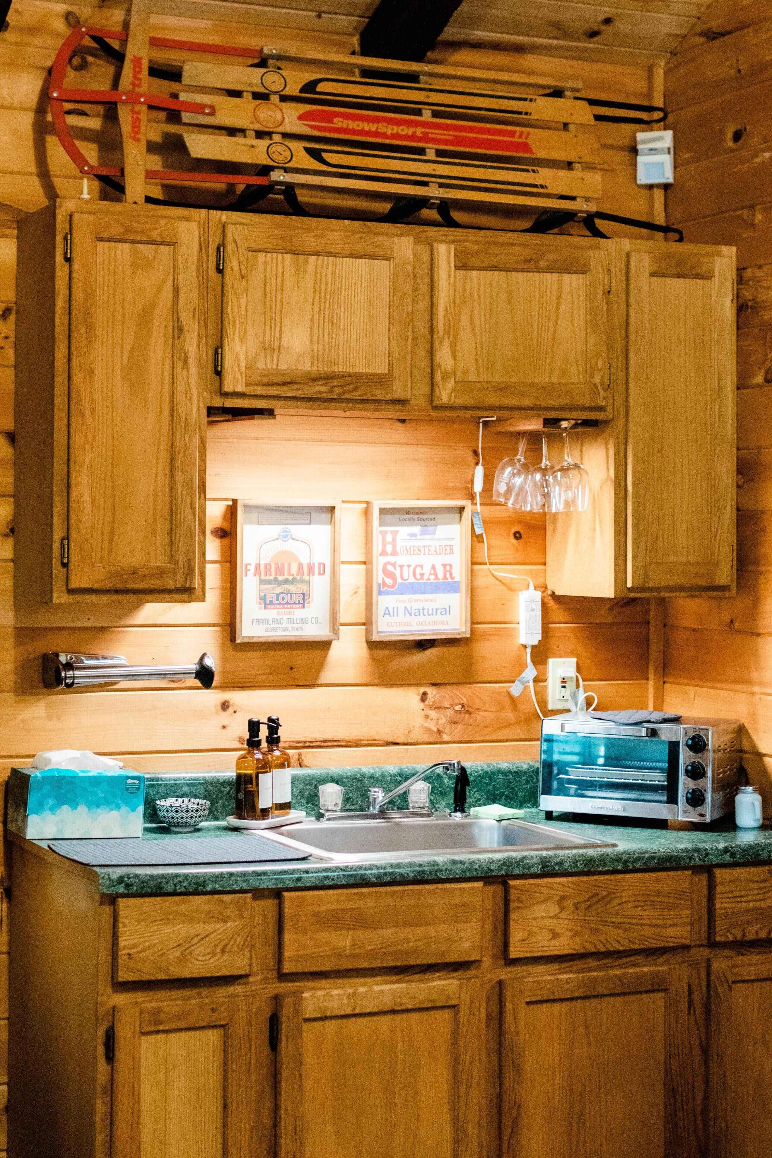 A wooden kitchen with a sink, countertop, and cabinets. There are framed signs on the wall, a toaster, and a bottle of hand sanitizer.