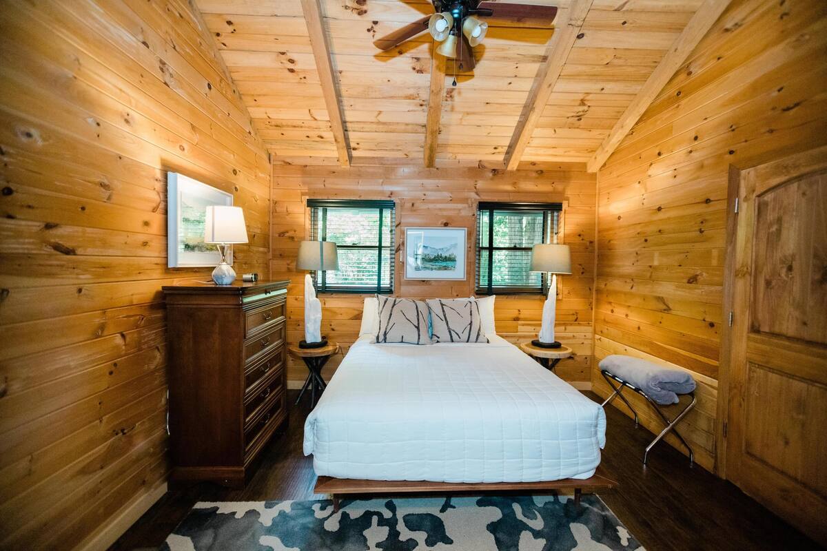 Un dormitorio de cabaña de madera con una cama, dos mesitas de noche con lámparas, un aparador y un ventilador de techo.