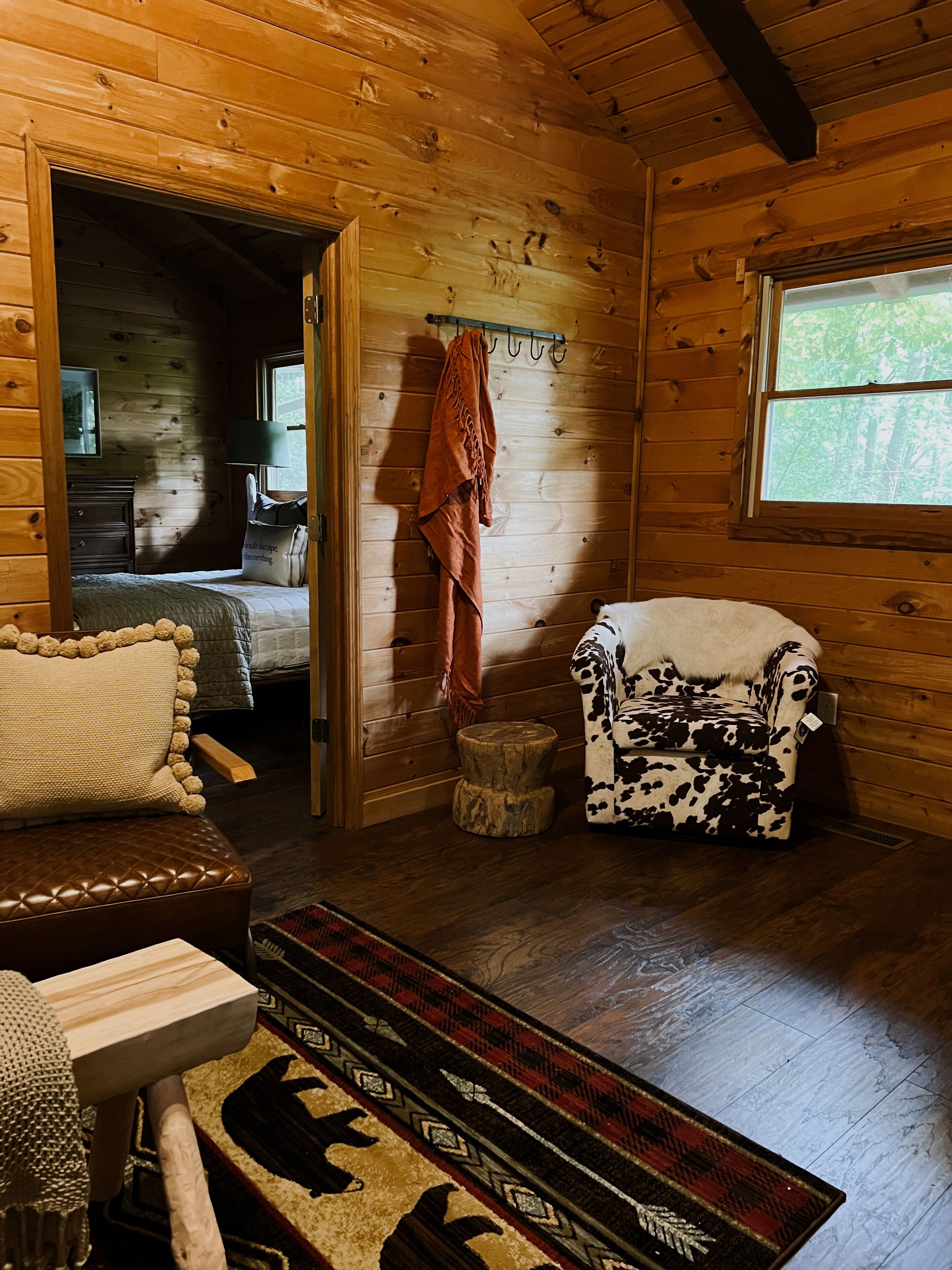 A cozy cabin interior with a bed, a wooden bench, a patterned rug, and a cow-print armchair.