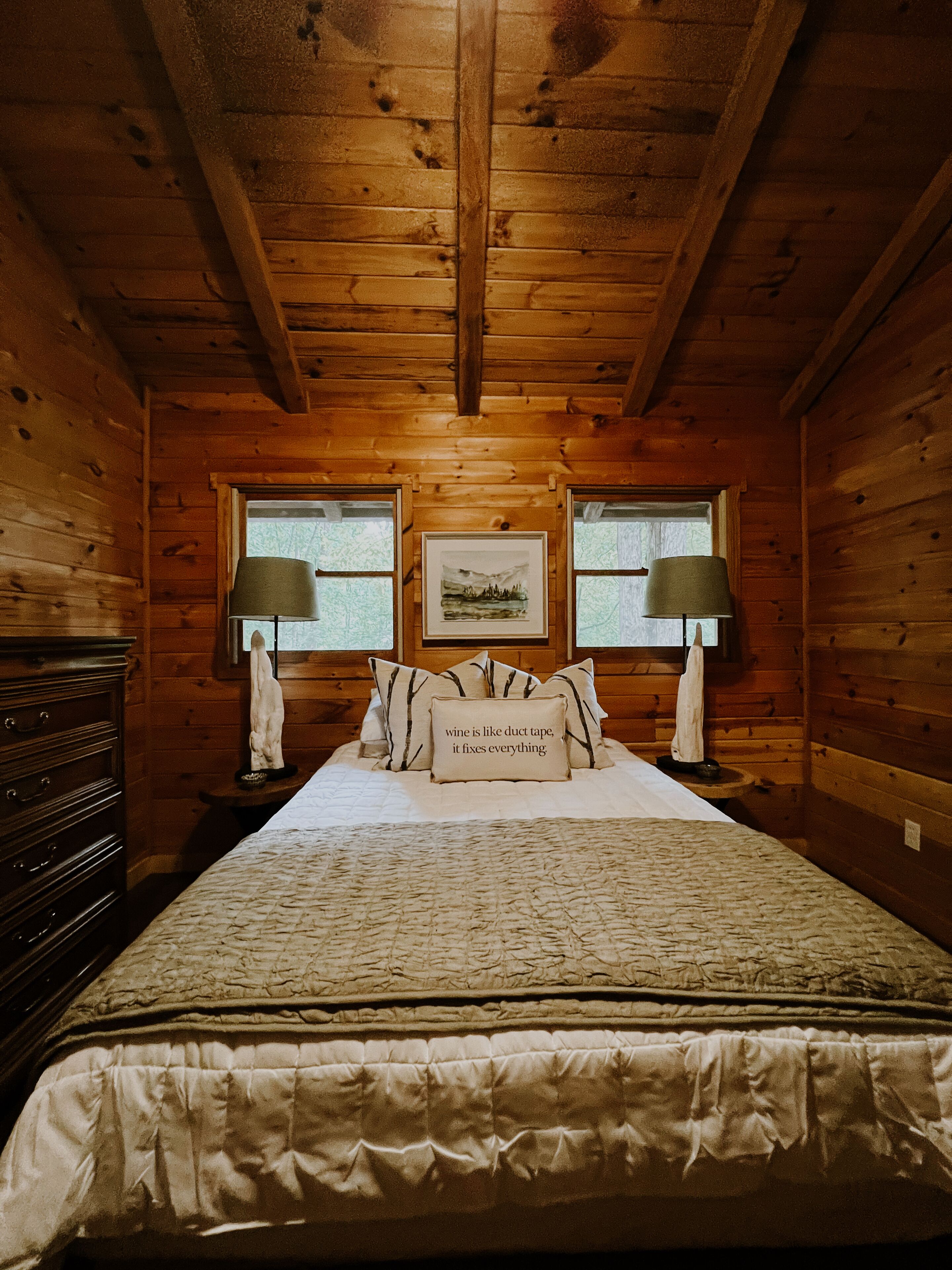 Une chambre à coucher dans une cabane en bois, avec un lit, deux lampes, une commode et un tableau encadré au mur.