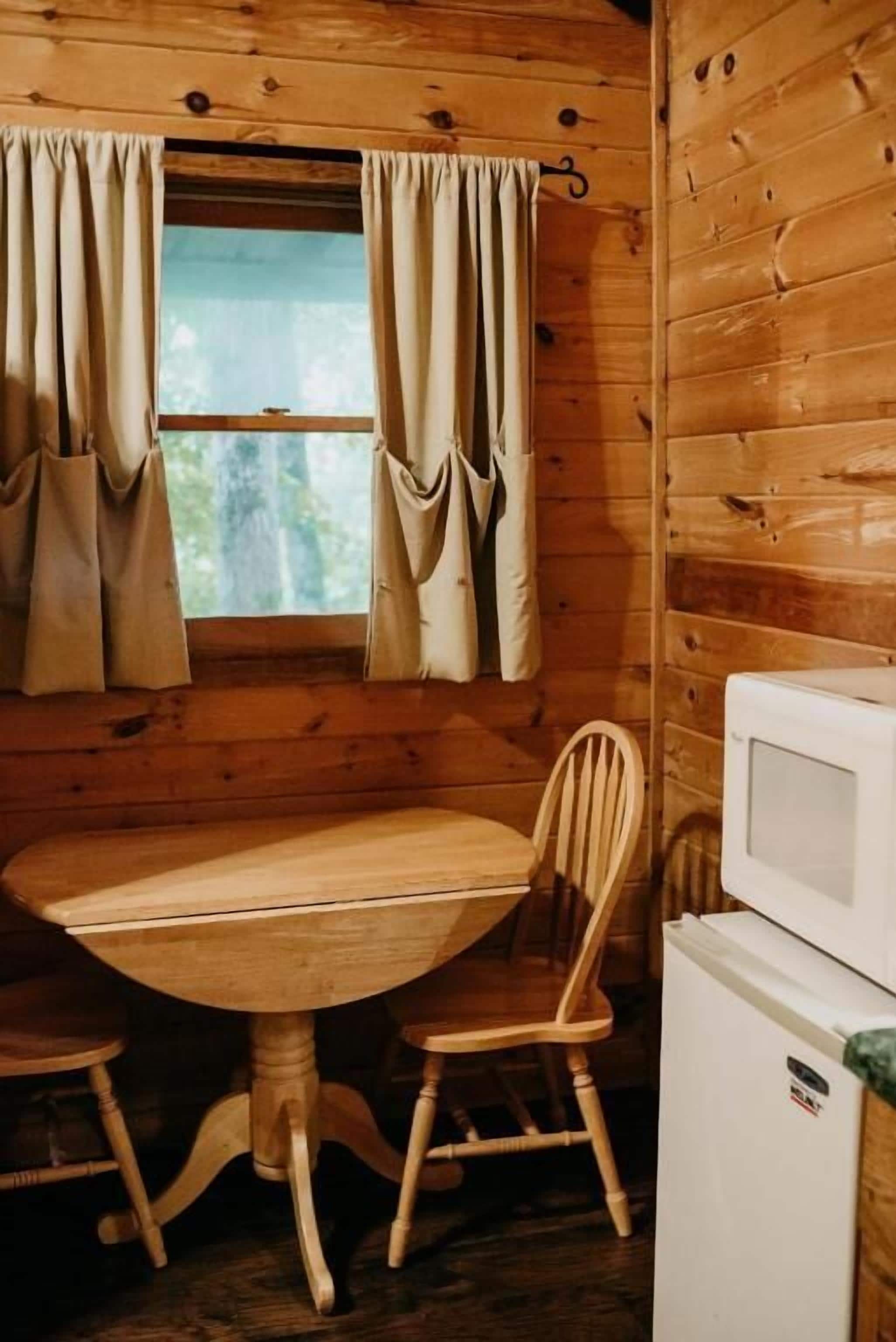 A wooden cabin interior with a dining table, two chairs, a window with curtains, and a white refrigerator.