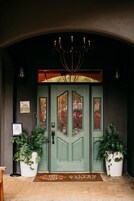 A green double door with stained glass panels, flanked by two potted plants, under a dark archway with a chandelier.