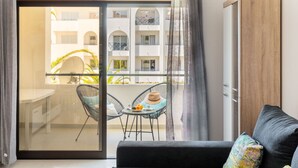 A modern living room with a sliding glass door leading to a balcony. The room features a black sofa, a small round table with a hat and fruit, and two wicker chairs. - Lovely apartment for 4 guests with pool, A/C, WIFI, TV and terrace (Armação De Pêra)