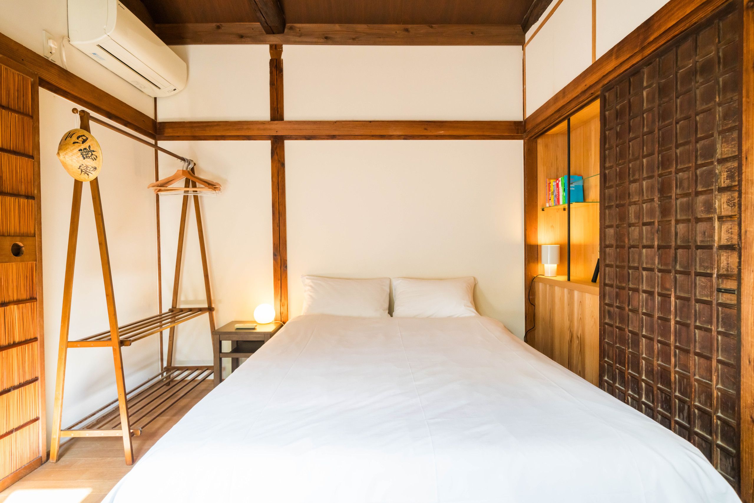 A bedroom with a bed, wooden beams, a hanging clothes rack, and a wall-mounted shelf with books.