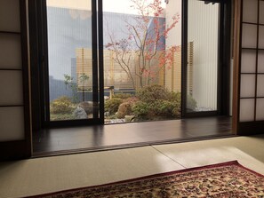 Une pièce de style japonais traditionnel, avec un sol recouvert de tatamis, un paravent en papier de riz (shoji) et une vue sur un jardin agrémenté de rochers et d’un arbre.