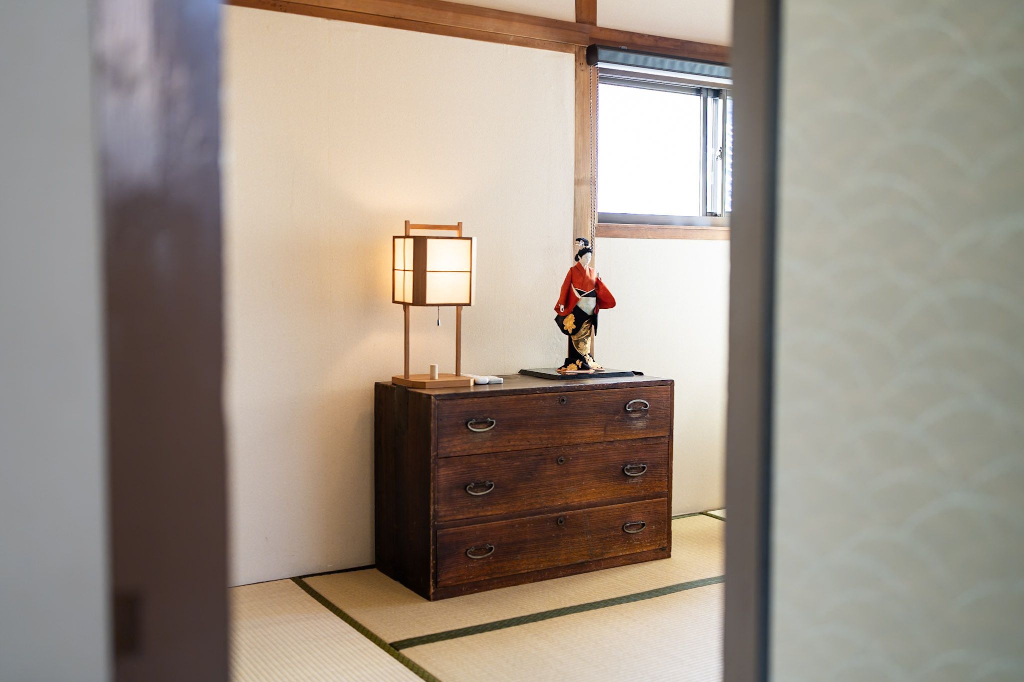 A traditional Japanese room with tatami flooring, a wooden dresser with drawers, a hanging lamp, and a decorative figure.