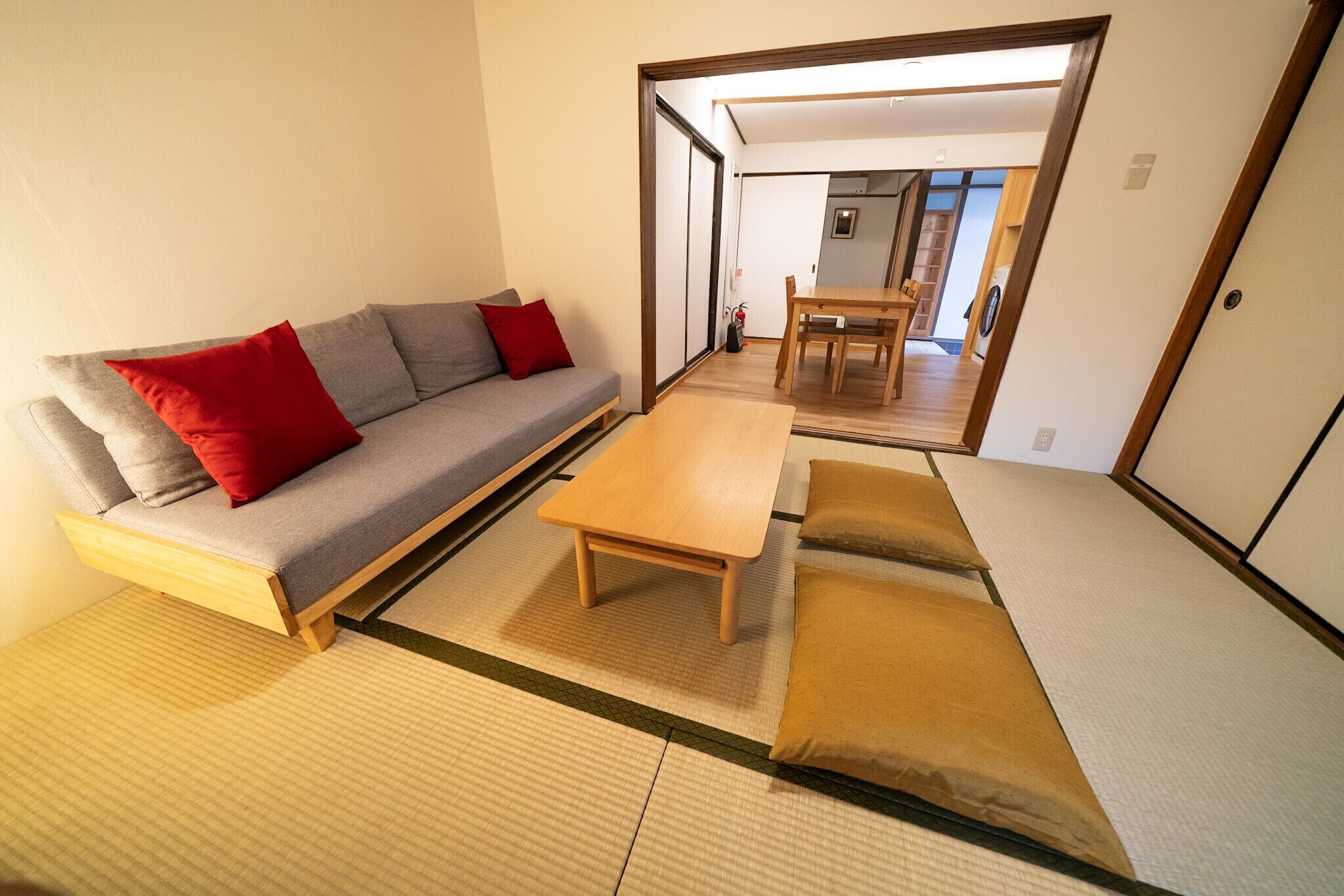 Una habitación tradicional japonesa con suelo de tatami, una mesa de madera baja, un sofá con cojines rojos y un comedor con mesa y sillas de madera.