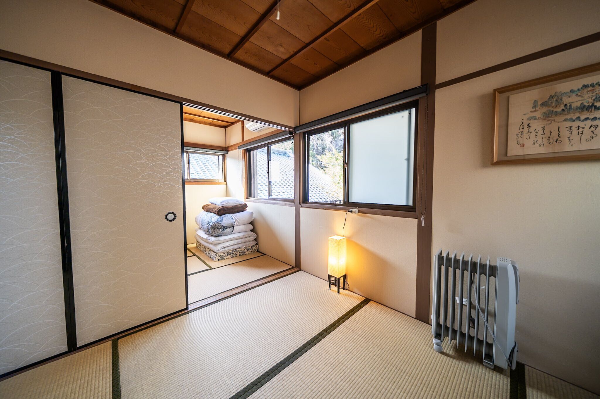 A traditional Japanese room with tatami flooring, a futon bed, a radiator, and a framed calligraphy piece on the wall.