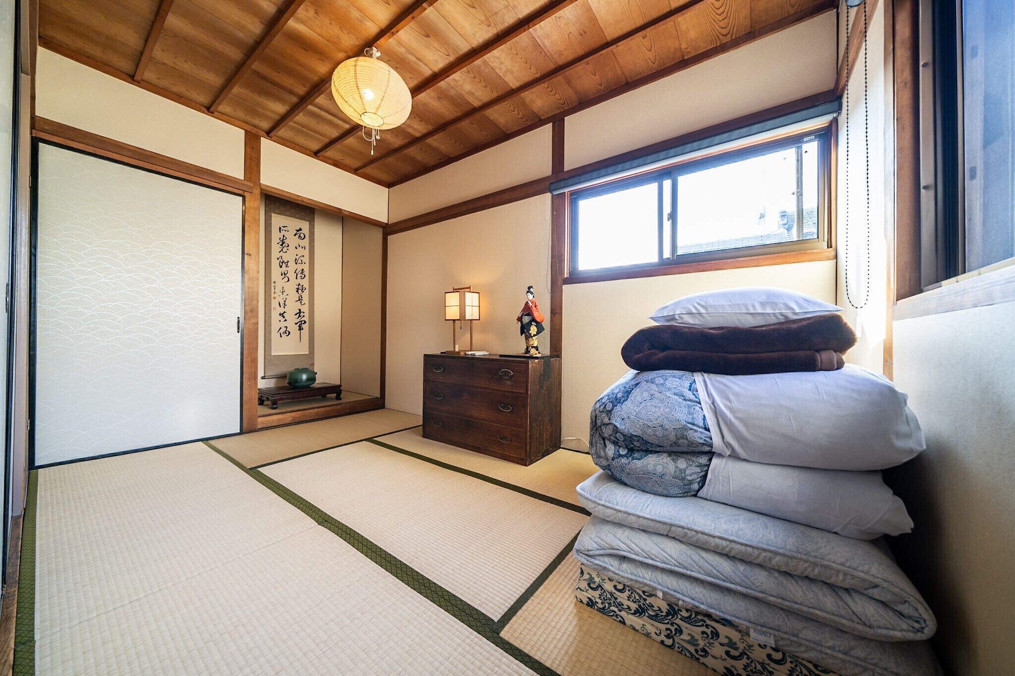 A traditional Japanese room with tatami flooring, a shoji screen, a wooden dresser, and a stack of futons.