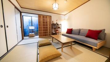 A traditional Japanese-style room with tatami flooring, a low wooden table, a sofa with cushions, a sliding door, and a view of a wooden fence outside.