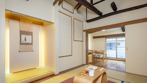 Une pièce de style japonais traditionnel, avec un sol recouvert de tatamis, une basse table en bois sur laquelle se trouve un plateau de fruits et une théière, et une porte coulissante donnant sur un espace repas.