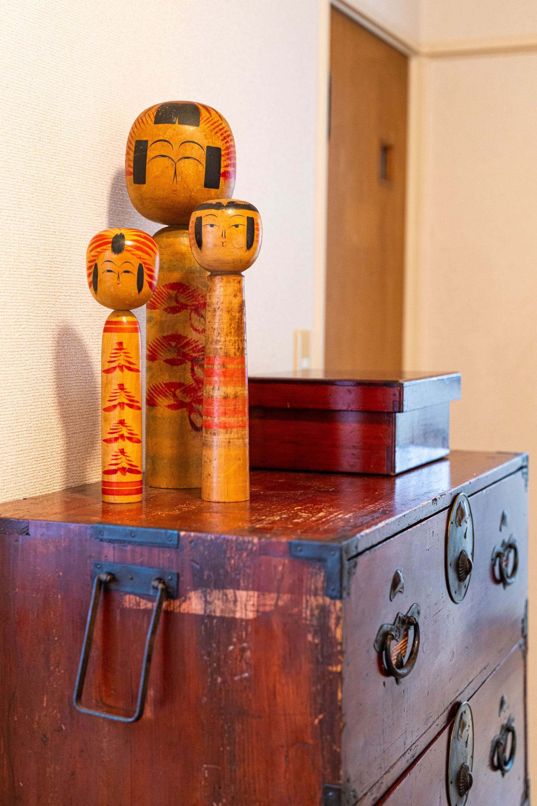 A wooden chest with metal handles and a latch, containing a red box and three wooden figures with painted faces.