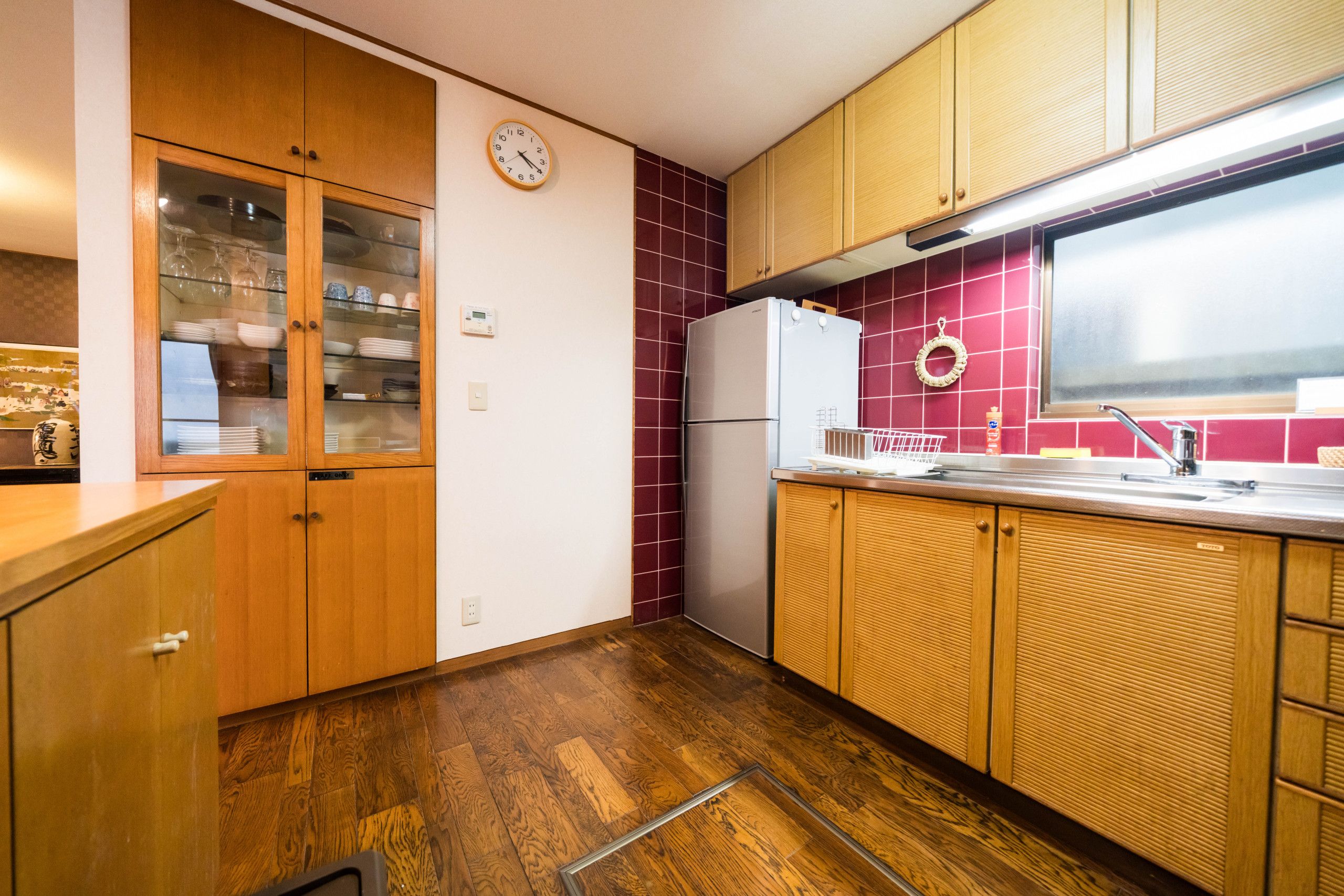 A kitchen with wooden cabinets, a refrigerator, a clock, and a tiled backsplash.
