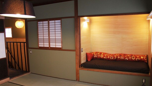 Une pièce traditionnelle japonaise avec un sol recouvert de tatamis, un lit bas en bois agrémenté de coussins à motifs rouges, une porte coulissante de type shoji et une lampe suspendue.
