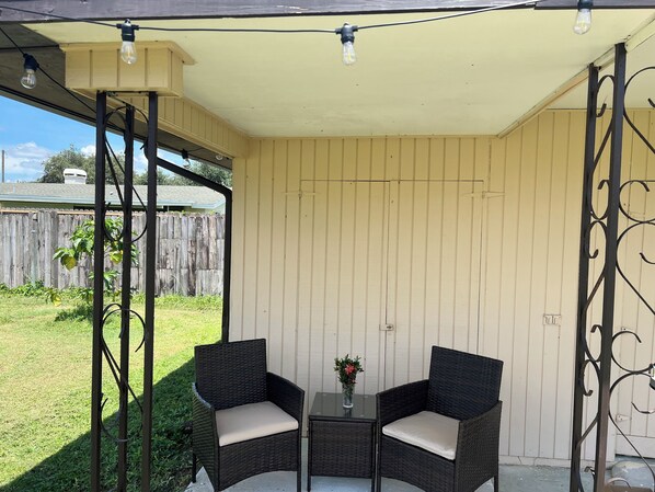A covered patio with two wicker chairs and a small table, a wooden fence in the background, and a clear sky.