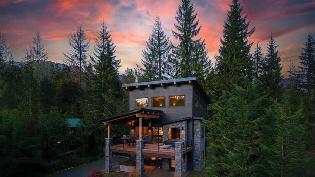 A modern two-story cabin with a deck, surrounded by dense forest.
