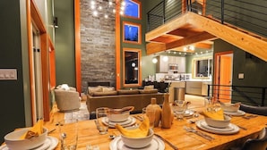A dining area with a wooden table set for four, a kitchen in the background, and a staircase leading to an upper level.